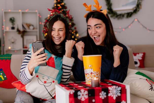 joyful pretty young girls keep fists and look at phone sitting on armchairs and enjoying christmas time at home