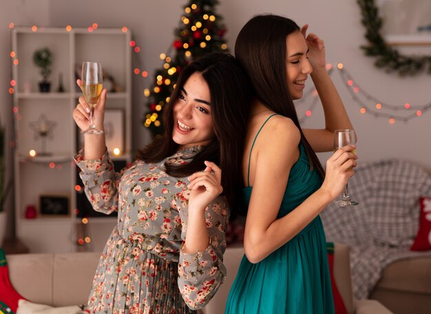 Joyful pretty young girls stand back to back holding glasses of champagne enjoying christmas time at home