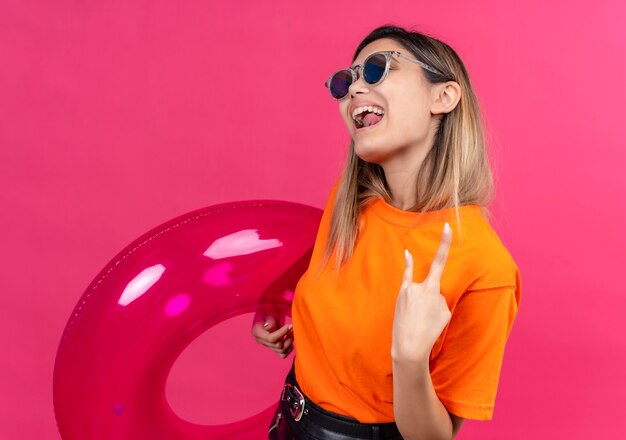 Радостная симпатичная молодая женщина в оранжевой футболке в солнечных очках демонстрирует рок-жест, держа розовое надувное кольцо на розовой стене