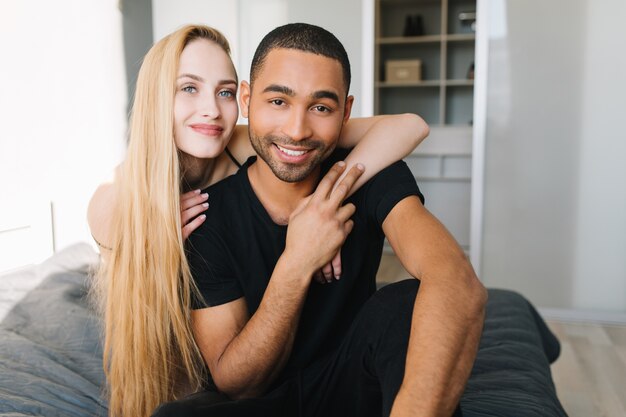 Joyful pretty young woman with long blonde hair hugging handsome guy from back. Couple in love chilling on bed at home in modern apartment, true emotions, lovely