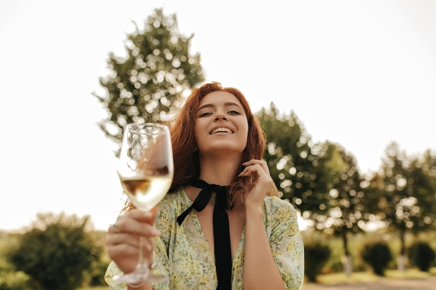 Free photo joyful red haired lady with black bandage on her neck in modern green dress smiling holding glass with drink and looking into camera outdoor