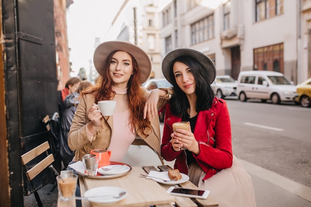 Joyful red-haired woman drinking tea while talking with friend