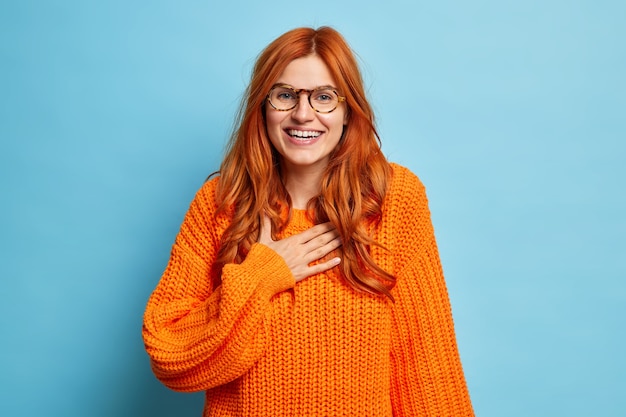 Free photo joyful red haired young woman giggles positively as hears funny news keeps hand on chest feels very happy wears glasses and knitted orange jumper.