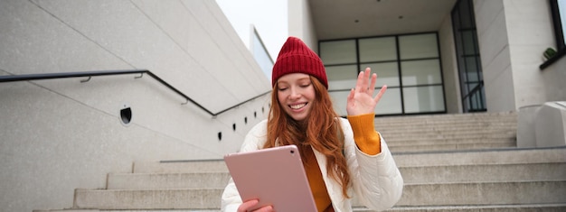 Joyful redhead teen girl student with digital tablet says hello waves hand at gadget camera connects