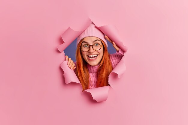 Joyful redhead young woman in eyewear laughs happily has joyful mood wears hat and optical glasses breaks through paper