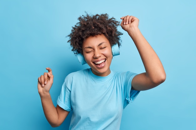 Joyful relaxed African American girl enjoys favorite playlist listens music via wreless headphones raises arms dressed casually isolated over blue wall. People hobby and lifestyle concept