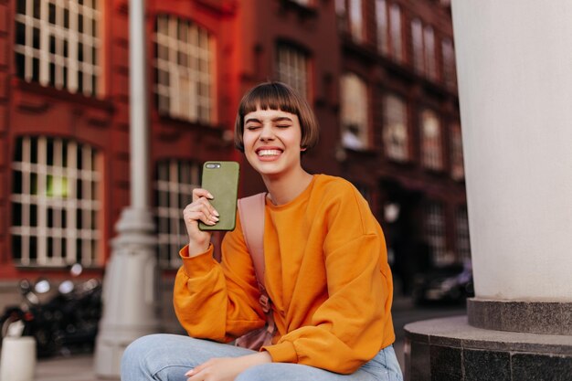 주황색 스웨트셔츠를 입은 즐거운 단발머리 여성은 밖에서 눈을 감고 미소 짓고 있습니다. Happy girl은 카키색 케이스에 스마트폰을 들고 있습니다