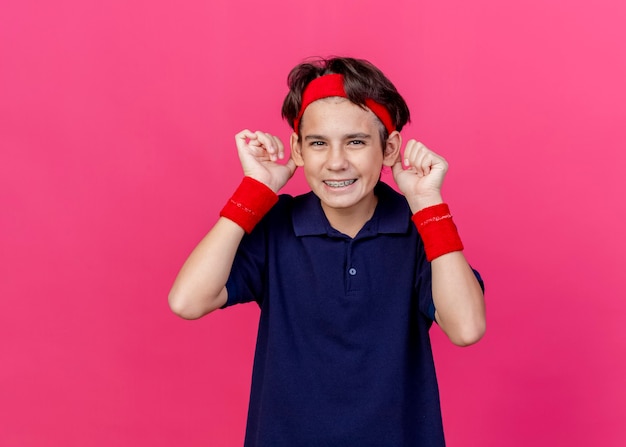 Gioioso sciocco giovane ragazzo sportivo bello che indossa la fascia e braccialetti con bretelle dentali che guarda l'obbiettivo che fa le orecchie di scimmia isolate su fondo cremisi con lo spazio della copia