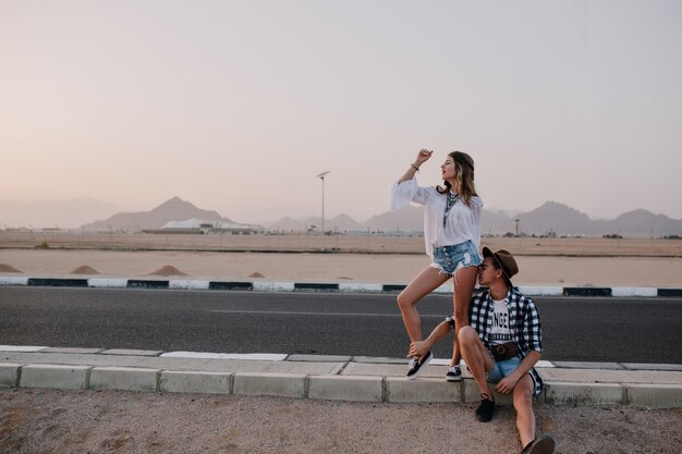 Joyful slim woman funny dancing while her tired boyfriend resting on the road on mountain. Portrait of adorable young woman and man traveling around country and waiting for a ride on highway