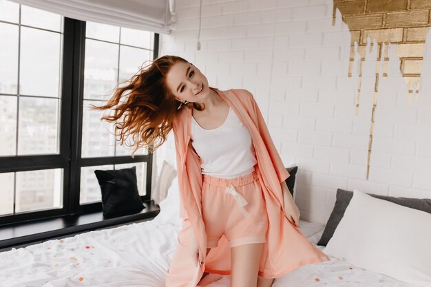 Joyful smiling girl posing on bed. Lovable european female model in pyjama relaxing in morning.