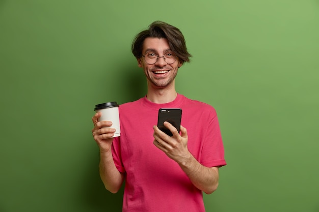 즐거운 웃는 남자는 온라인 상점에서 필요한 것을 검색하고, 스마트 폰 애플리케이션을 사용하고, 소셜 네트워크를 탐색하고, 종이컵에서 향기로운 커피를 마시고, 트렌디 한 헤어 스타일을 가지고, 실내에서 포즈를 취합니다.