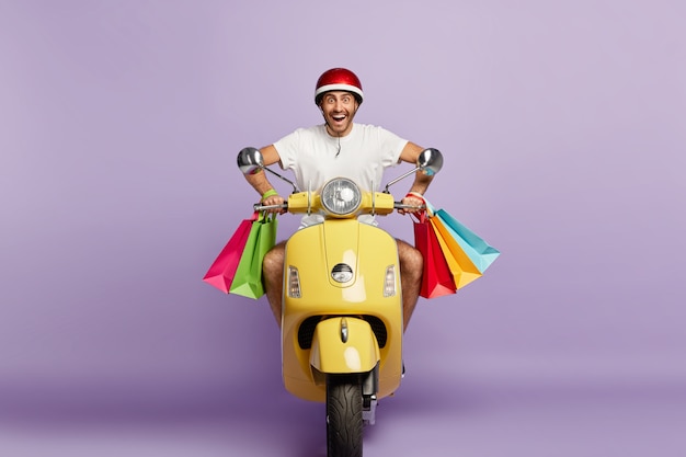 Free photo joyful smiling guy with helmet and shopping bags driving yellow scooter