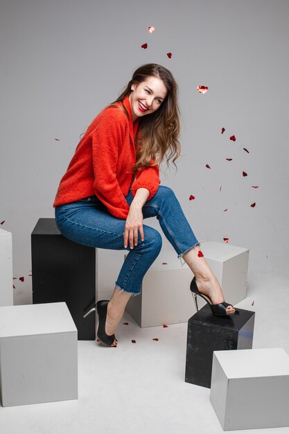 Joyful smiling young woman under festive confetti wearing red sweater, jeans posing on black and white cubes in studio