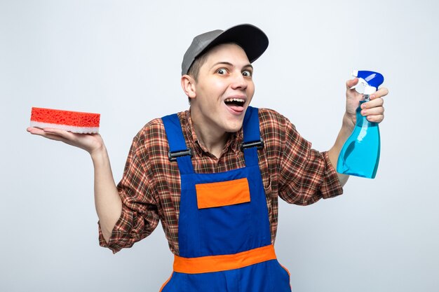 Joyful spreading hand young cleaning guy wearing uniform and cap holding sponge with cleaning agent 