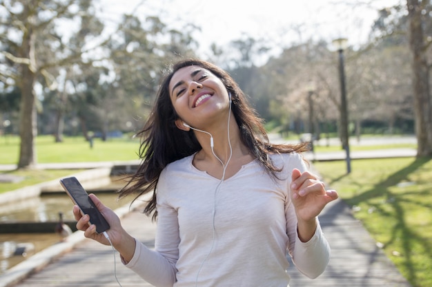 Joyful student girl in earphones holding smartphone and dancing