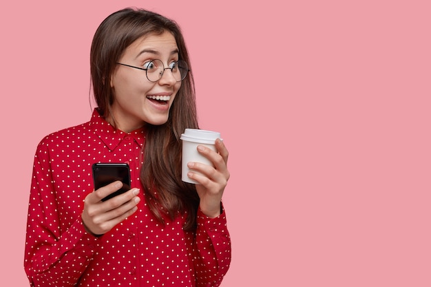 Joyful student with long black hair, notices funny scene aside, holds modern cell phone and disposable cup of coffee, wears glasses