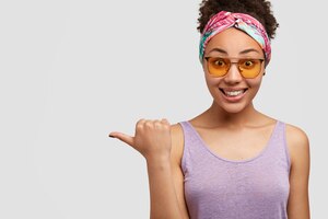 Free photo joyful stylish black lady wears colourful headband, yellow shades and purple vest, indicates aside, glad to advertise new item in shop, rejoices discounts, isolated over white wall, copy space