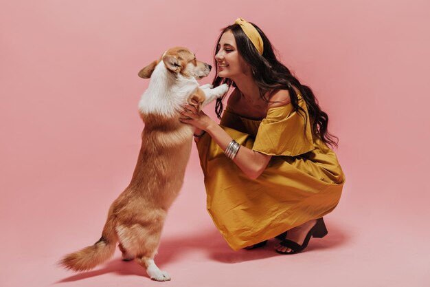 Joyful stylish girl with black wavy hair in bandana and long yellow sundress looking at corgi and playing with her on isolated background