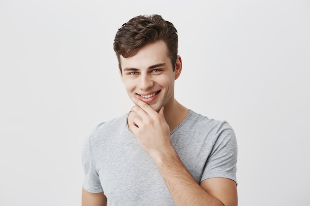 Joyful stylish handsome caucasian male in gray t-shirt keeps hand under chin, smiles gently, happy or pleased to hear compliments, feels proud of himself, has positive expression