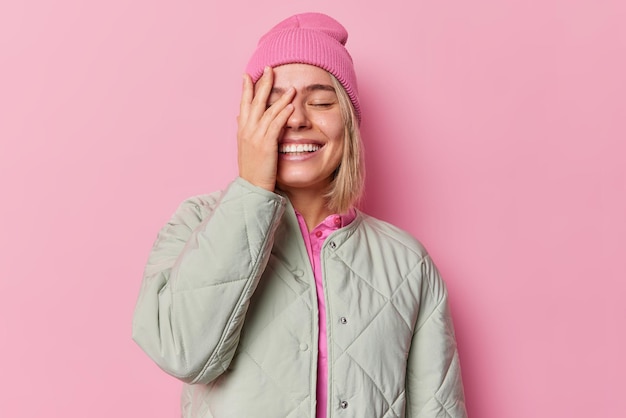 Joyful stylish millennial girl covers face with palm giggles positively shows white teeth dressed in jacket wears hat feels happy expresses sincere authentic emotions isolated over pink background.