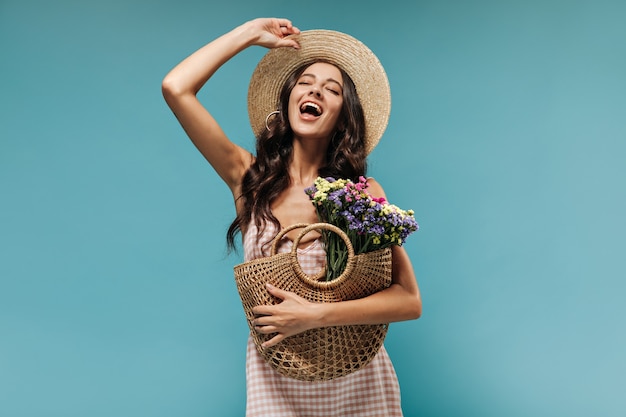 Free photo joyful stylish woman with long curly hair in modern hat and plaid clothes screams and posing with straw bag and colorful flowers