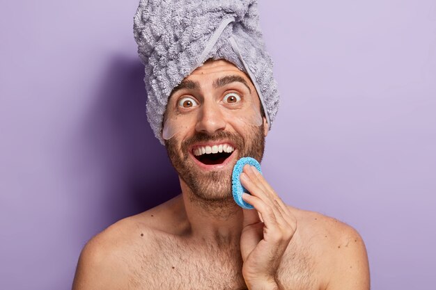 Joyful surprised man wants to have smooth skin, wipes face with cosmetic sponge, applies silicone patches under eyes