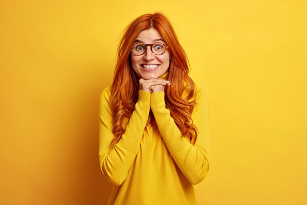 Joyful surprised redhead young woman keeps hands under chin shows white teeth feels glad listens with great interest interlocutor dressed in casual jumper.