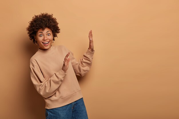 Joyful surprised woman raises palms towards brown space, steps aside from something