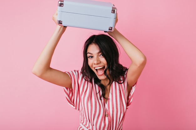 Joyful tanned girl with cheerful mood is dancing with briefcase. Lady in striped top smiles broadly.