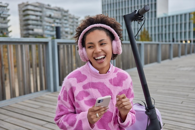 Joyful teenage girl enjoys favorite audio track in headphones rests after riding electric scooter poses at bridge outside against urba setting wears casual jumper. Youth hobby and lifestyle concept