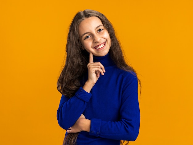 Joyful teenage girl looking at camera touching cheek with finger isolated on orange wall with copy space