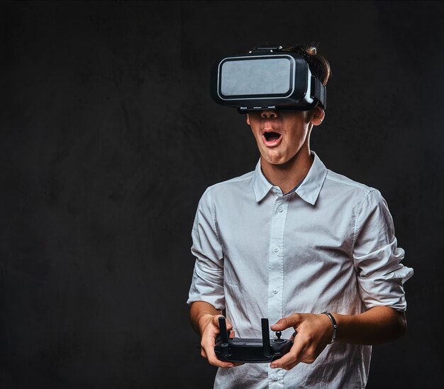 Joyful teenager dressed in a white shirt wears virtual reality glasses and controls the quadcopter using the control remote. Isolated on the dark background.