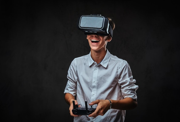 Joyful teenager dressed in a white shirt wears virtual reality glasses and controls the quadcopter using the control remote. Isolated on the dark background.