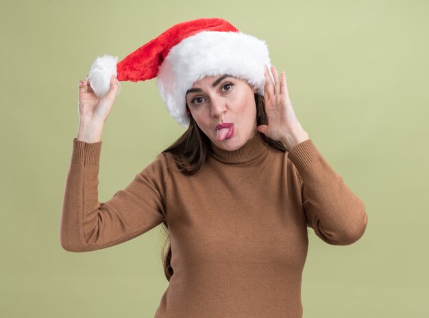 オリーブグリーンの壁に分離された舌を示すクリスマス帽子をかぶってうれしそうな傾斜頭の若い美しい少女