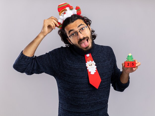 白い背景で隔離の舌を示すクリスマスのおもちゃを保持している髪のフープとクリスマスのネクタイを身に着けているうれしそうな傾いた頭の若いハンサムな男