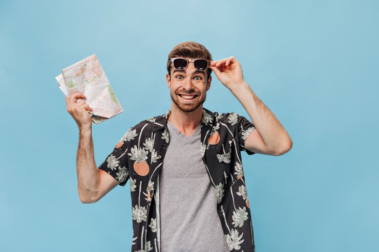 Free Photo joyful trendy tourist in grey t-shirt, printed shirt and sunglasses looking at front and holding map on isolated wall