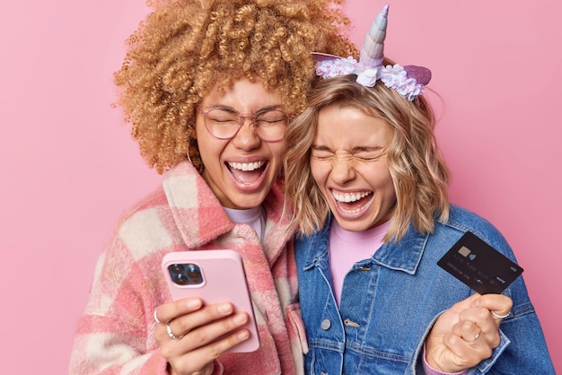 Радостные две подруги громко восклицают, одетые в модную одежду, держат мобильный телефон и кредитную банковскую карту, совершают покупки в Интернете или легко переводят деньги, изолированные на розовом фоне студии