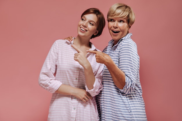 Joyful two ladies with short hair in striped pink and blue summer clothes smiling and showing to place for text on isolated backdrop. 