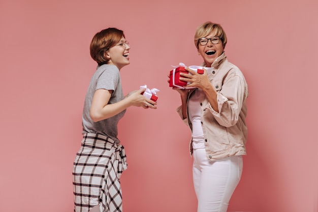 Joyful two short haired ladies with cool glasses in stylish clothes laughing and holding red gift boxes on pink background.