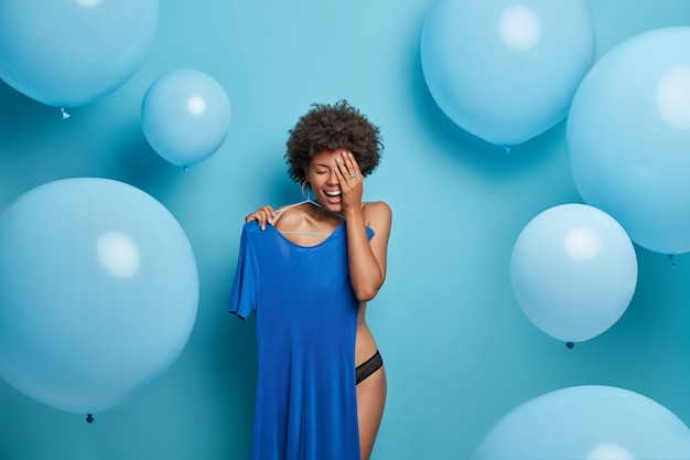 Joyful undressed Afro American young woman holds dress on hangers, dresses for blue theme wedding celebration, laughs positively, poses against blue wall, big balloons. Monochrome shot