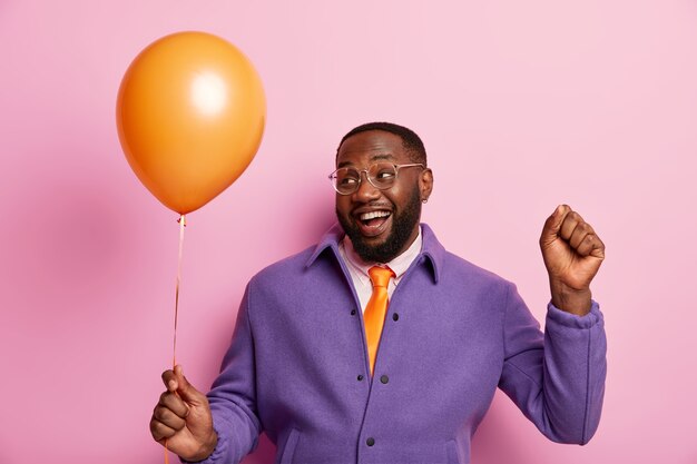 Joyful unshaven plump man celebrates something, has playful mood, raises fist, holds inflated balloon