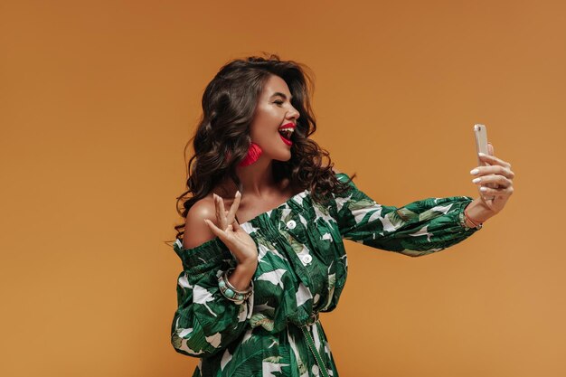Joyful wavy haired woman with red lips and tanned skin in printed green dress with belt makes photo and laughing on orange backdrop