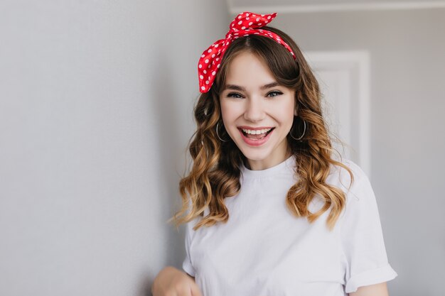 Joyful white girl posing with excited face expression. Indoor photo of blithesome female model with red ribbon in curly hair.
