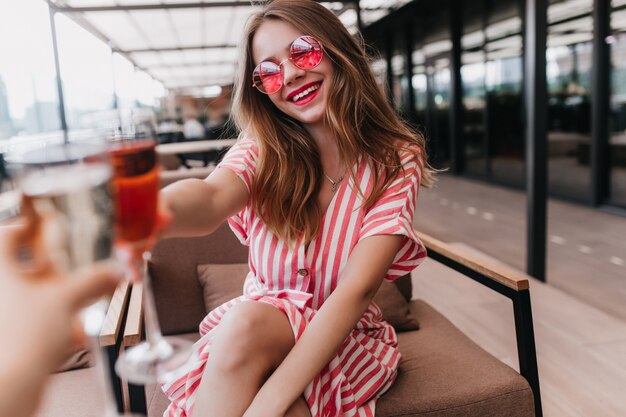 Радостная белая девушка в летнем платье проводит время в кафе. Портрет чувственной блондинки в розовых очках, выражающей счастье в теплый день.