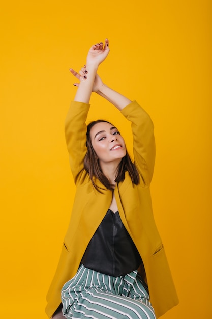 Joyful white woman in elegant yellow jacket posing with hands up. indoor photo of playful brunette caucasian girl