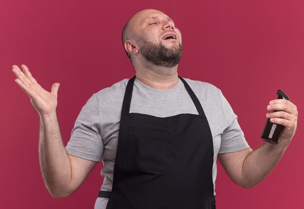 Joyful with closed eyes middle-aged male barber in uniform holding spray bottle spreading hand isolated on pink wall