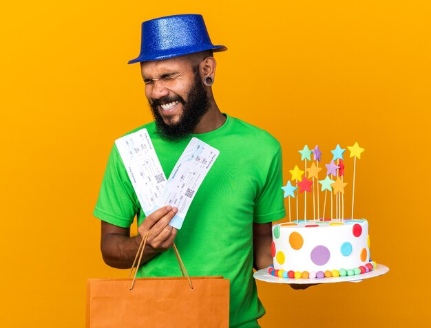 Joyful with closed eyes young afro-american guy wearing party hat holding gift bag and cake with tickets 