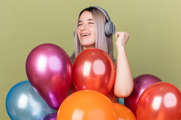 Free photo joyful with closed eyes young beautiful girl wearing dental braces with headphones standing behind balloons