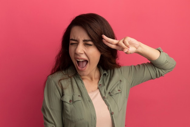 Joyful with closed eyes young beautiful girl wearing olive green t-shirt putting fingers on temple isolated on pink wall