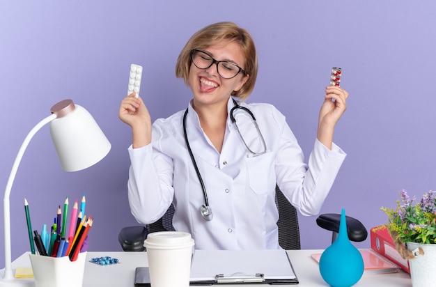 Joyful with closed eyes young female doctor wearing medical robe with stethoscope and glasses sits at table with medical tools holding pills and showing tongue isolated on blue wall